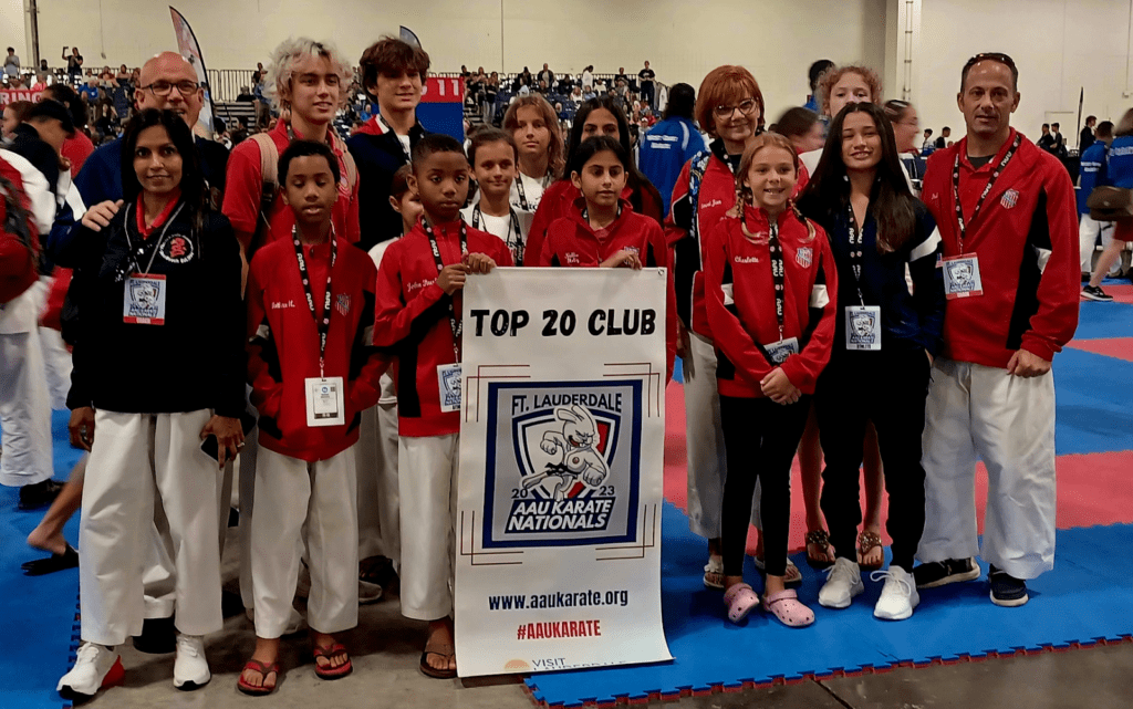 Youth karate team in red jackets holding a “Top 20 Club” sign at a competition.