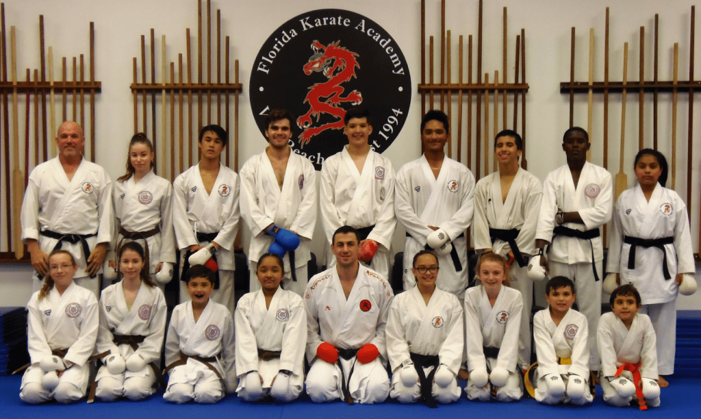Group of karate students and instructors posing in a dojo with training weapons on the wall.