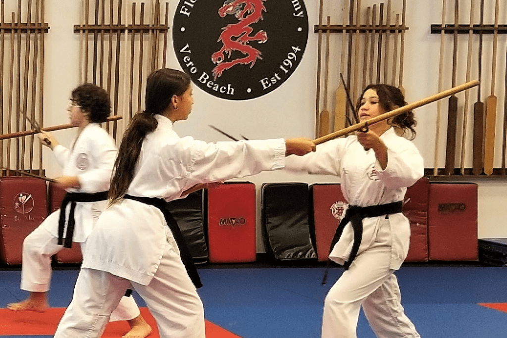 Students in karate uniforms practice bo staff sparring inside a dojo, with training weapons and pads on the wall behind them.
